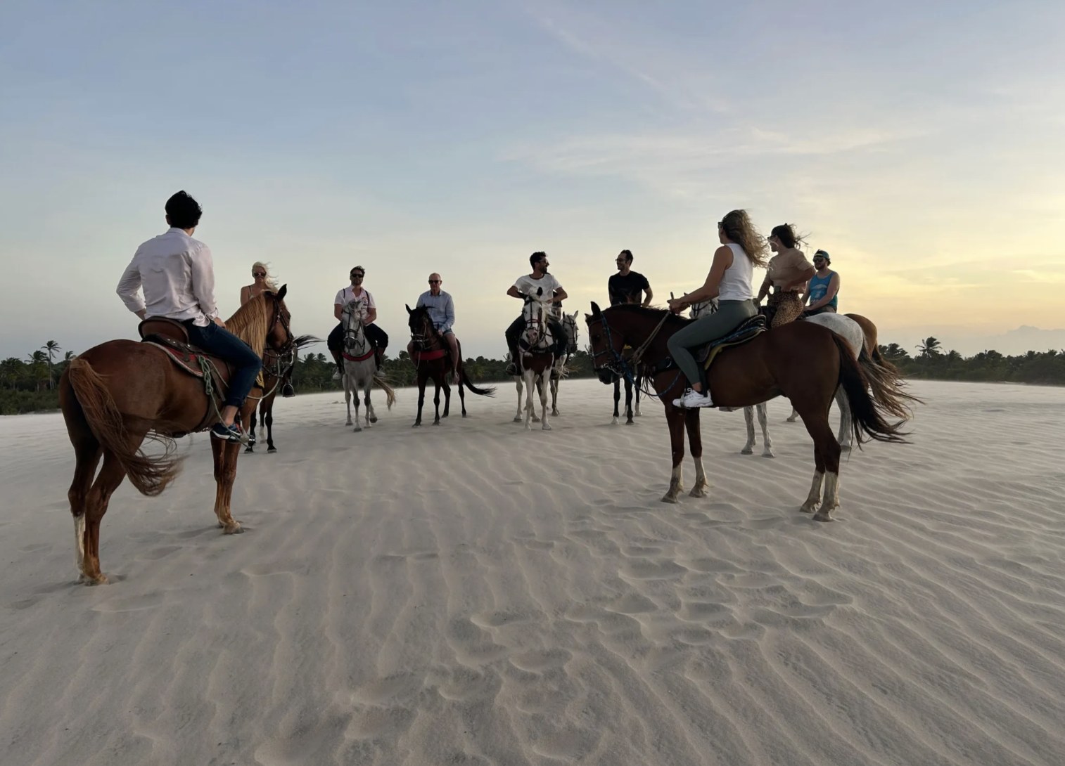 The crew riding horses across white sand dunes at sunset